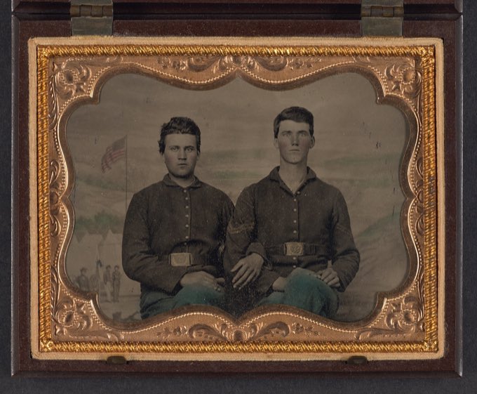 Hand-colored tintype of two unidentified Union soldiers seated before a painted studio backdrop of a military camp; the backdrop scenery is outlined, not the faces.